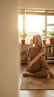 Mature Caucasian woman practicing yoga meditation at home in warm sunlight with houseplants on windowsill creating peaceful wellness atmosphere.