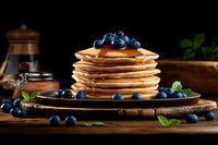 Stack of golden pancakes topped with fresh blueberries and maple syrup on rustic wooden board, dark moody background with vintage kitchenware and mint leaves.