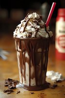 Decadent chocolate milkshake topped with whipped cream, chocolate shavings and drizzle served in tall glass with red straw on wooden surface. Dark chocolate pieces scattered.