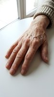 Senior man's weathered hand resting on white surface near window, wearing beige knit sweater. Depicts aging, wrinkles, and life experience for healthcare and wellness content.