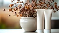 White cosmetic tubes near ceramic vase with dried flowers on table. Minimalist beauty product mockup with natural organic decoration and soft lighting.