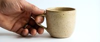 Hand holding a rustic speckled ceramic mug on white background, showcasing handmade pottery craft for kitchen and lifestyle concepts.