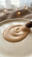 Paintbrush applying swirled beige paint on ceramic plate with warm bokeh lights and fabric backdrop, suitable for art and craft projects.