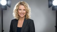 Professional blonde businesswoman smiling confidently in dark suit jacket against studio backdrop with bright lighting for corporate headshots.