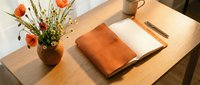 Cozy workspace with open leather journal, ceramic vase with orange flowers, coffee mug and pen on wooden desk creating warm productive atmosphere.