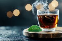 Steaming hot tea in glass cup on wooden coaster with fresh green leaf and bokeh lights in dark background creates cozy atmosphere for relaxation.