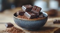 Dark chocolate pieces and cocoa powder in rustic ceramic bowl on wooden surface, moody food photography with selective focus and natural lighting.