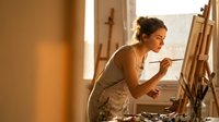 Young mixed-race woman artist painting on canvas in warm sunlit studio workspace with easel and art supplies for creative hobby and artistic expression.