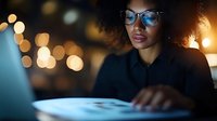 Young African American businesswoman wearing glasses working late on laptop computer in modern office with bokeh city lights background at night.