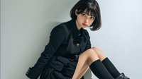 Young Asian woman in dark green military style jacket sitting on floor wearing black knee-high socks against neutral background for fashion lifestyle content.