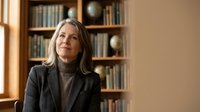 Middle-aged woman with shoulder-length gray hair, Caucasian ethnicity, wearing black blazer and turtleneck, sitting in front of wooden bookshelf. Professional portrait for business, corporate, and leadership concepts.