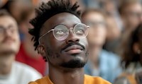 Young African American man with glasses and natural hair looking up thoughtfully, wearing orange sweater against blurred crowd background. Authentic emotional portrait.