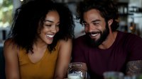Happy mixed-race couple enjoying wine together at home, smiling and laughing in cozy kitchen setting, romantic date night atmosphere for lifestyle content.