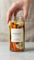 Caucasian hand holding glass jar filled with mixed pickled vegetables including cauliflower, carrots, and peppers with artisan label on neutral background for food packaging and culinary content.