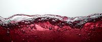 Red wine or grape juice with bubbles and foam splashing against glass surface, creating dynamic wave pattern against white background.