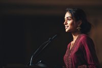 Young South Asian woman in burgundy dress speaking at podium with microphone during professional conference or business presentation event.