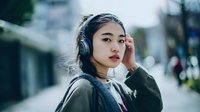 Young Asian woman wearing headphones listening to music on urban street with soft bokeh background for lifestyle and technology concepts.