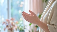 Woman hands in prayer position near window with soft natural light and flowers creating peaceful meditation atmosphere for wellness and spirituality.