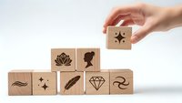 Hand placing wooden cube with star symbol on pyramid of blocks featuring luxury icons including diamond, rose, feather and silhouette on white background.