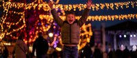 Young man celebrating at festive Christmas market with arms raised in joy surrounded by warm golden lights and holiday decorations during winter evening.