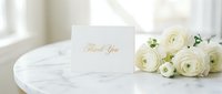 Elegant thank you card with gold script beside white ranunculus flowers on a marble table, soft natural light, wedding stationery concept.