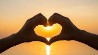 Hands forming heart shape silhouette against golden sunset sky over water creating romantic love symbol for Valentine's Day greeting cards.