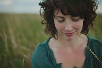 Young woman with dark curly hair and gentle smile looking down thoughtfully, wearing teal blouse against blurred natural grassland background in soft evening light.