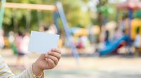 Child's hand holding blank white card at playground with blurred play equipment and children in background, suitable for advertising, promotions, messages, or educational content.
