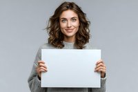 Young woman with curly brown hair wearing gray sweater holding blank white paper against neutral background, perfect for adding custom message.