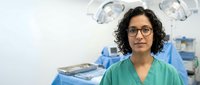 Female Hispanic surgeon with curly hair and glasses wearing green scrubs standing confidently in modern operating room with surgical lights and equipment.