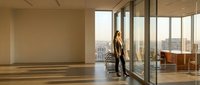 Professional businesswoman in elegant suit standing by floor-to-ceiling windows in modern office space overlooking urban cityscape at sunset.