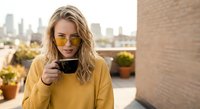Young blonde woman in yellow sweater enjoying coffee on urban rooftop terrace with city skyline background. Lifestyle portrait for morning routine concepts.