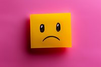 Yellow sticky note with hand drawn sad face emoji on vibrant pink background showing negative emotion, depression concept for mental health awareness.
