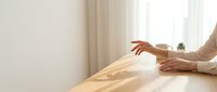Elegant female hands resting on a wooden table in a bright minimalist interior with soft natural light and sheer curtains.