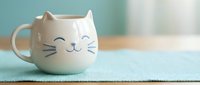Adorable white ceramic cat mug with cute face design sitting on turquoise placemat, perfect for coffee tea lovers and kitchen decor enthusiasts.