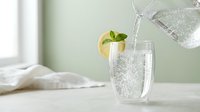Fresh sparkling water being poured into glass with lemon slice and mint garnish on kitchen counter for healthy hydration and refreshing drink concept.