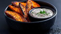 Roasted sweet potato wedges served with creamy herb dip in black ceramic bowl. Close up view of appetizing vegetarian dish on dark background.