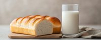 Fresh baked white bread loaf sliced on wooden cutting board with glass of milk against neutral background, minimalist food composition.