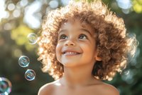 Mixed race child with curly afro hair smiling while watching soap bubbles float in sunlight against blurred natural background with bokeh effect.