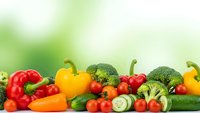 Fresh organic vegetables including bell peppers, broccoli, tomatoes, carrots and cucumbers arranged on white surface with green bokeh background for healthy eating.