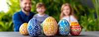 Colorful decorated Easter eggs with traditional folk patterns in blue, orange and yellow colors displayed on table with blurred family background.