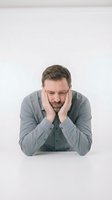 Stressed middle-aged Caucasian businessman in gray shirt holding head in hands showing fatigue, burnout and mental exhaustion at white desk.