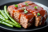 Sliced homemade meatloaf with rich tomato glaze sauce served on black plate with steamed green asparagus spears and fresh herb garnish, comfort food dinner.