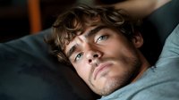 Young Caucasian man with blue eyes and curly brown hair lying on couch in moody lighting, looking up pensively with intense gaze and slight stubble on face.