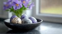 Decorative speckled Easter eggs in black ceramic bowl near window with purple spring flowers, moody natural lighting creates seasonal ambiance.