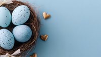 Speckled blue Easter eggs in natural twig nest with golden heart decorations on pastel blue background. Minimal spring holiday composition with copy space.