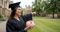 Happy young Caucasian female graduate in cap and gown holding piggy bank on university campus lawn, celebrating education investment and financial planning.