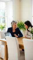 Senior businesswoman mentoring young female colleague at modern office table with laptop discussing career development and professional growth strategies.