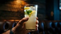 Caucasian male hand holding a tall glass of lemonade with ice, lemon slices, and fresh mint in a dark bar setting.