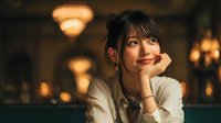 Young Asian woman with bangs resting chin on hand in warm cafe lighting, smiling thoughtfully at camera with cozy restaurant atmosphere and bokeh lights.